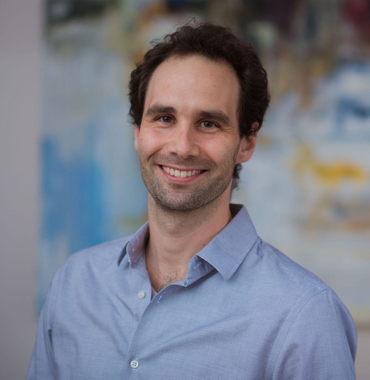 The researcher Siniša Hrvatin wears a blue shirt and smiles at the camera. He stands in front of a painting.