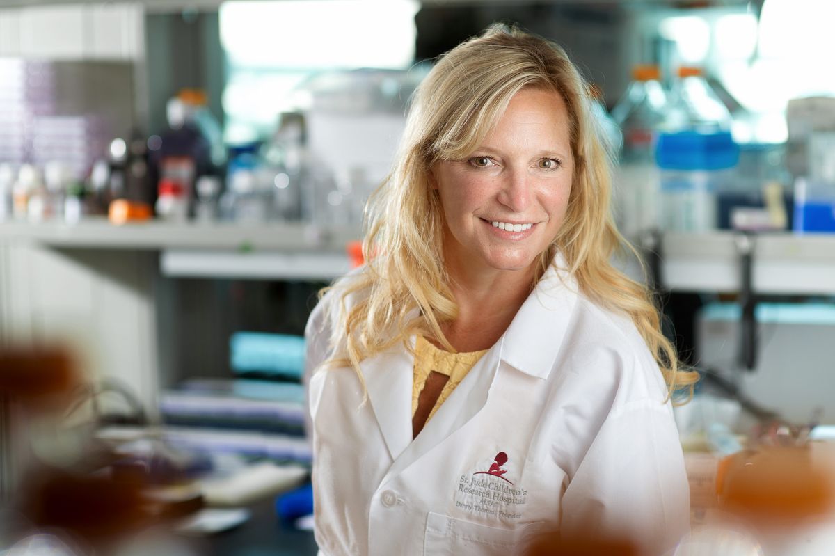 Stacey Schultz-Cherry wears a white lab coat and stands in a laboratory full of research equipment.