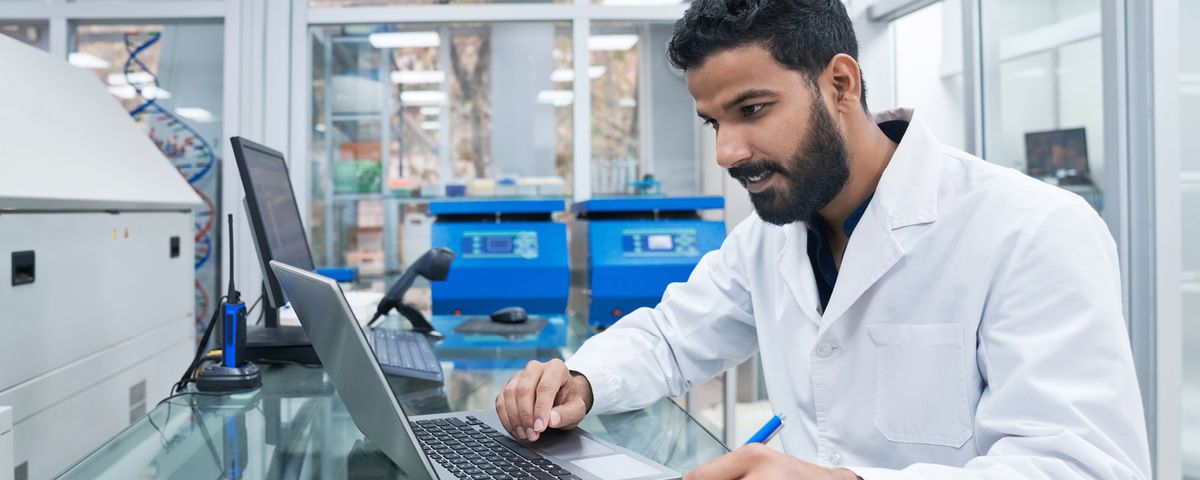 A person sitting at a laboratory bench, typing on a laptop while looking at notes on a clipboard.