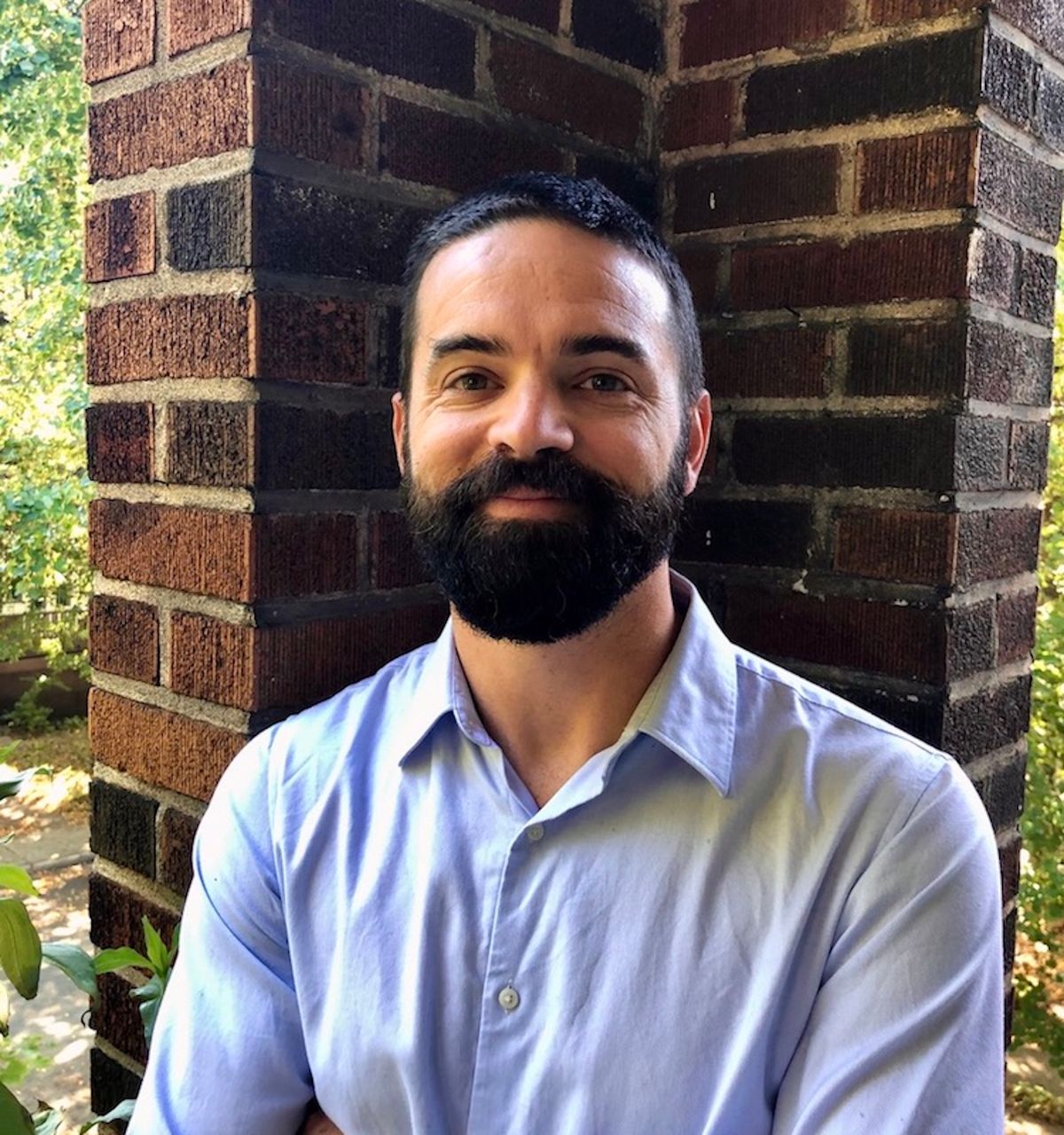 Matthew Harrington wears a blue collared shirt and stands outside in front of a brick wall.