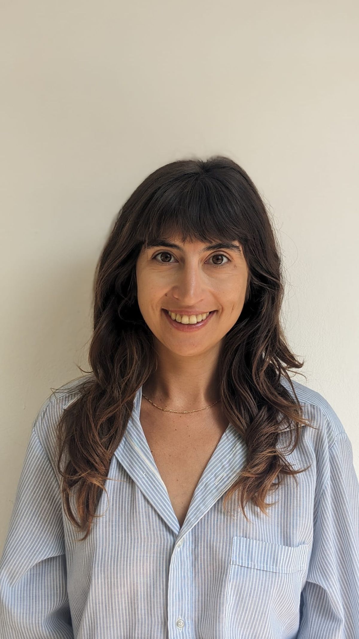 Hedva Haykin wears a blue button-down shirt and smiles in front of a white wall.