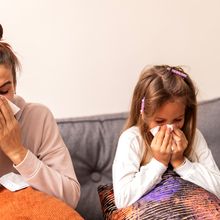 A woman and a child sitting on a couch blowing their noses.&nbsp;