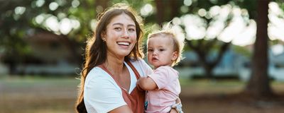 A smiling woman holds a young child outside in the park.