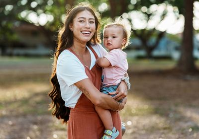 A smiling woman holds a young child outside in the park.