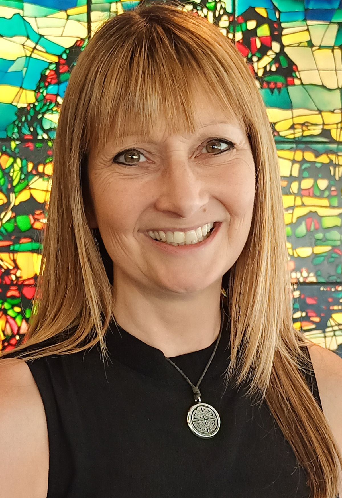 A woman wears a black sleeveless shirt and smiles at the camera. She stands in front of a stained-glass window.