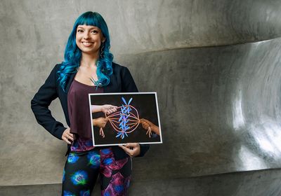 Beata Mierzwa, a postdoctoral researcher at University of California San Diego and the founder of Beata Science Art, poses for a photograph holding a print of one of her science illustrations. She has bright blue hair and is wearing leggings from her microscopy print design that features blue and green and red and blue cells.