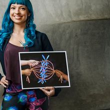 Beata Mierzwa, a postdoctoral researcher at University of California San Diego and the founder of Beata Science Art, poses for a photograph holding a print of one of her science illustrations. She has bright blue hair and is wearing leggings from her microscopy print design that features blue and green and red and blue cells.