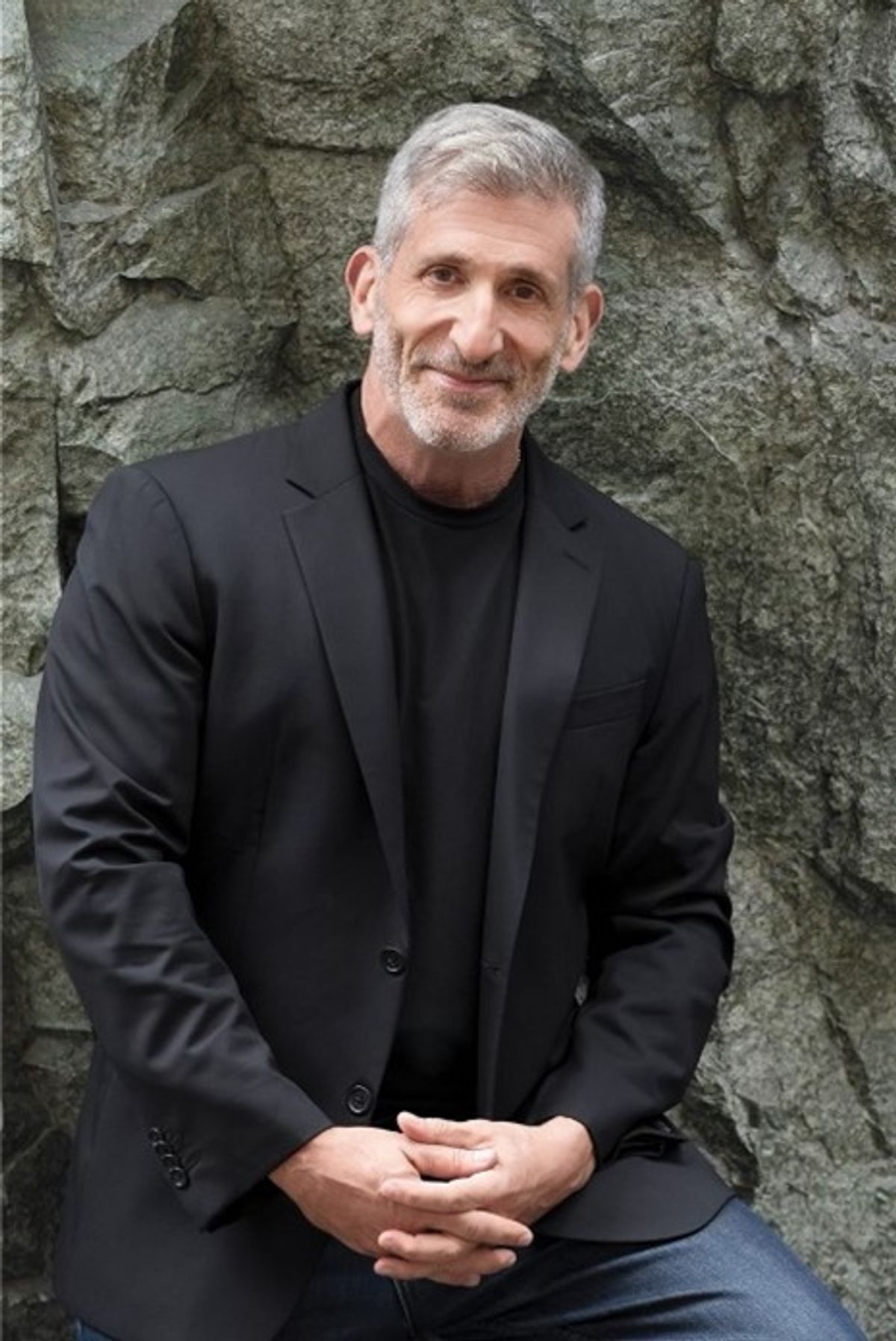 Photo of a scientist with interlaced fingers, wearing a black shirt and blazer, leaning against a rocky backdrop outdoors.