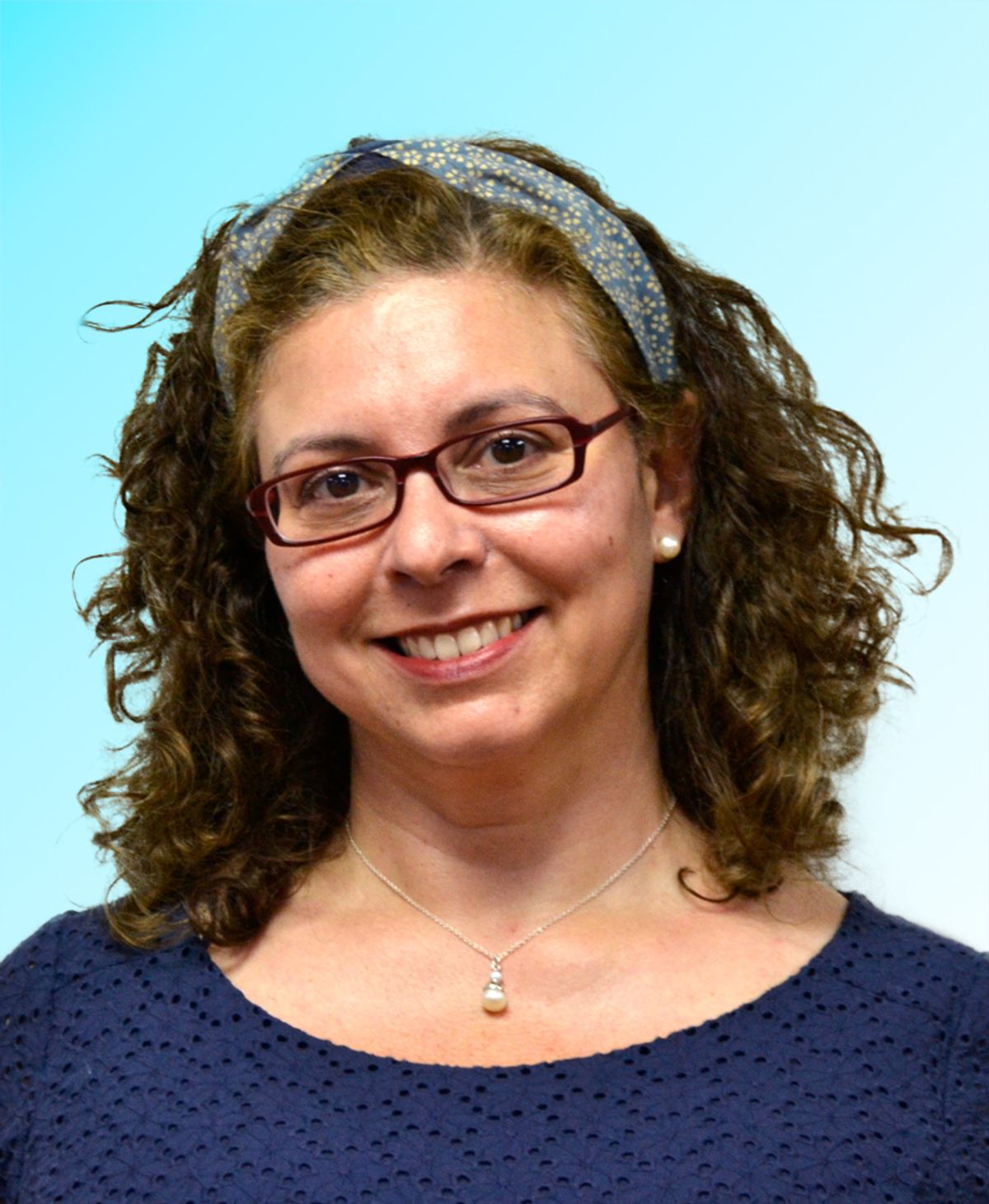 A woman with wavy hair wears a blue shirt and glasses. She smiles at the camera.  