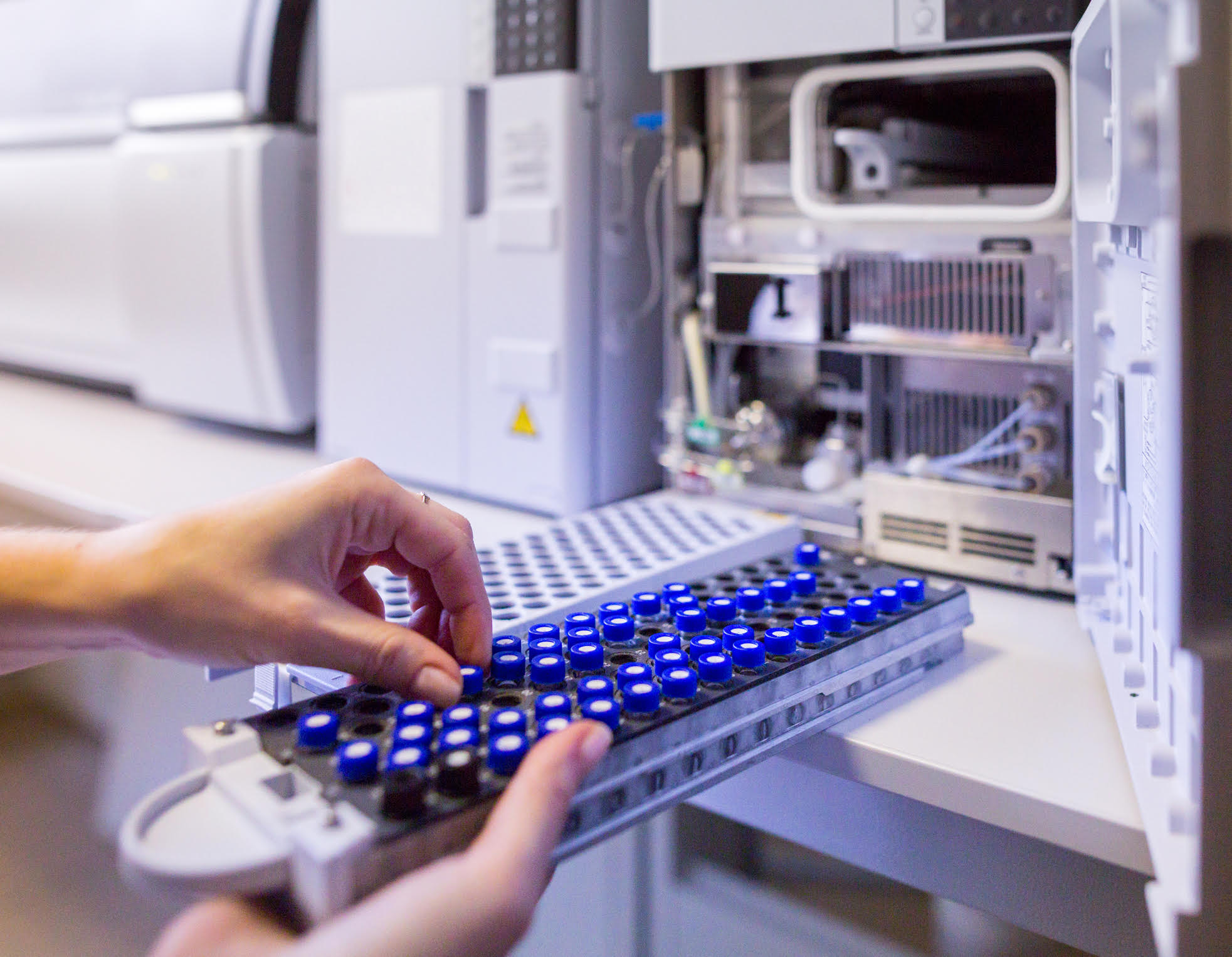 A scientist takes a small vial with a blue lid from a rack of similar vials, ready to place it in a liquid-chromatography mass spectrometer (LC-MS).