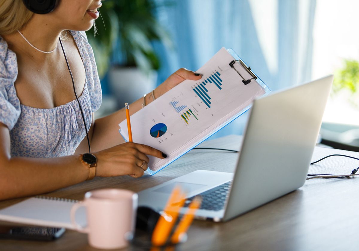 An individual looking at graphs and charts on a clipboard in front of a laptop.