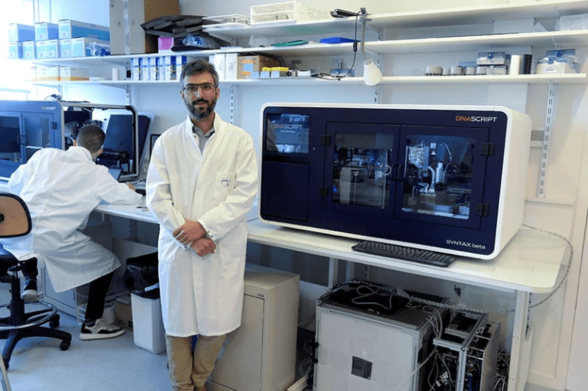 Photo of Thomas Ybert wearing a lab coat and standing next to a DNA Script benchtop DNA printer. 